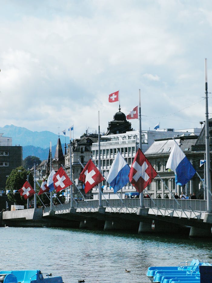 Picturesque view of Swiss flags on a bridge in Zurich with a backdrop of buildings and mountains.